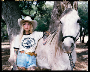Blonde woman in western wear standing next to a white horse.