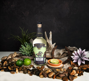 Image of a bottle of Ranch Rider Spirits Lime-infused Blanco Tequila with a blue, lime green, and gold label. At the top of the label, the Ranch Rider Spirits Co. logo and an illustration of a cowboy riding a horse. The label says “Lime Tequila, Blanco tequila infused with real fruit and all natural flavors. 60 Proof, 750 ml, 30% ABV.” The bottle is surrounded with rocks, driftwood, and limes.