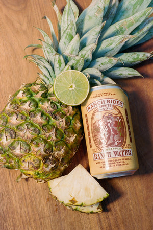 An image of the Pineapple Ranch Water pre-made cocktail can. The can is light yellow with brown, cream, and green details. The label says "A premium tequila seltzer" at the top. Below that, there is an image of a cowboy on a horse with a rope on a desert backdrop. Below the logo, the label says: "Pineapple Ranch Water, with only reposado tequila, sparkling water, real lime and pineapple." The can is resting against a whole pineapple on a wooden background with pineapple and lime slices.