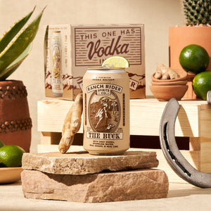An image of the Ranch Rider The Buck pre-made cocktail can. The can is light khaki with brown, yellow, and cream details. The label says "A premium vodka seltzer" at the top. Below that, there is an image of a cowboy on a horse with a rope on a desert backdrop. Below the logo, the label says: "The Buck, with only vodka, sparkling water, real ginger and real lime." The can is surrounded with rocks, succulents, limes, ginger, and other western ephemera.