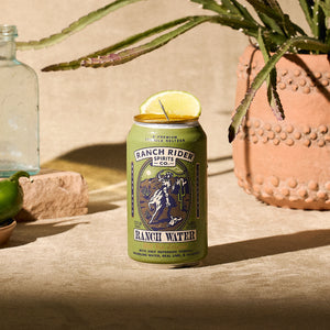 An image of the Jalapeño Ranch Water pre-made cocktail can. The can is olive green with dark blue, cream, and orange details. The label says "A premium tequila seltzer" at the top. Below that, there is an image of a cowboy on a horse with a rope on a desert backdrop. Below the logo, the label says: "Jalapeño Ranch Water, with only reposado tequila, sparkling water, real lime and jalapeño." The can is sitting in a desert backdrop with a succulent plant in the background. The can has a lime wedge on the rim.