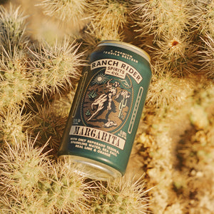 An image of the Ranch Rider Margarita pre-made cocktail can. The can is dark teal with dark blue, cream, light teal, and orange details. The label says "A premium tequila seltzer" at the top. Below that, there is an image of a cowboy on a horse with a rope on a desert backdrop. Below the logo, the label says: "Margarita, with only reposado tequila, sparkling water, sea salt, real lime and orange." The can is sitting on a cactus bush.